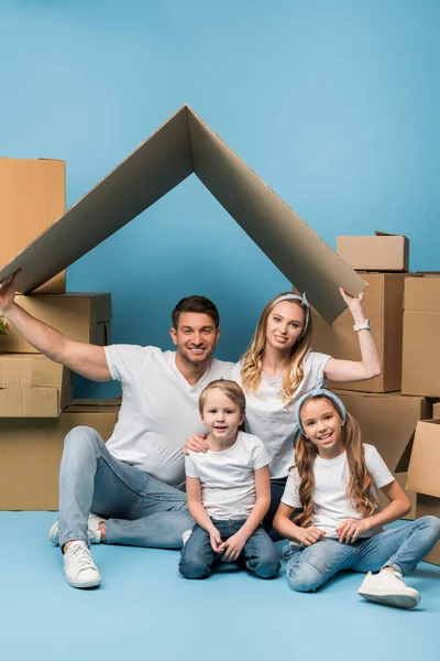 Positive parents holding carton roof over kids on blue with cardboard boxes for relocation — Stock Photo