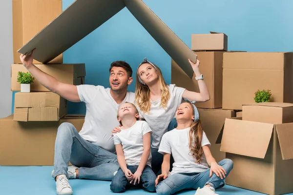 Happy parents holding carton roof over kids on blue with cardboard boxes for relocation — Stock Photo