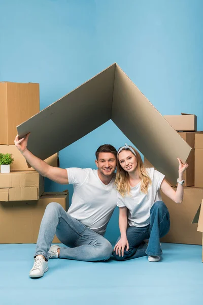 Alegre pareja sosteniendo techo de cartón sobre cabezas mientras está sentado en azul con cajas de cartón para la reubicación — Stock Photo