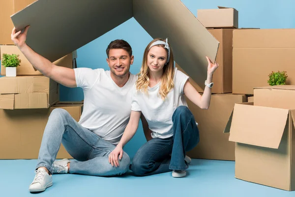 Beautiful couple holding carton roof over heads while sitting on blue with cardboard boxes for relocation — Stock Photo