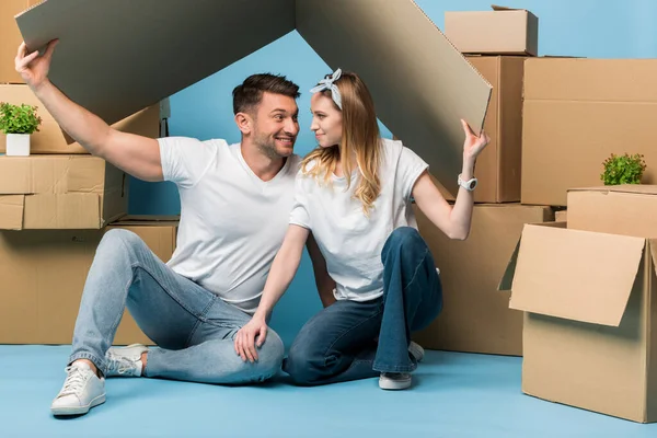 Счастливая пара, держащая картонную крышу над головой, сидя на синем фоне с картонными коробками для перемещения — стоковое фото
