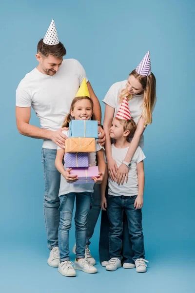 Familia feliz en gorras de fiesta de cumpleaños, hija sosteniendo cajas de regalo en azul - foto de stock