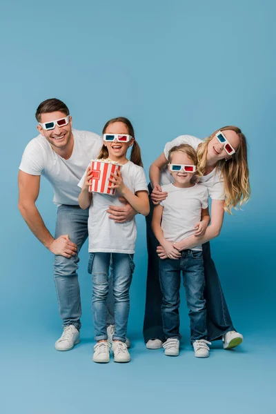 Gai famille regarder film en 3d lunettes et tenant seau de maïs soufflé sur bleu — Photo de stock