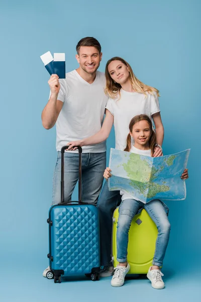 Sonrientes padres e hija con bolsas de viaje, mapa, boletos y pasaportes en azul - foto de stock