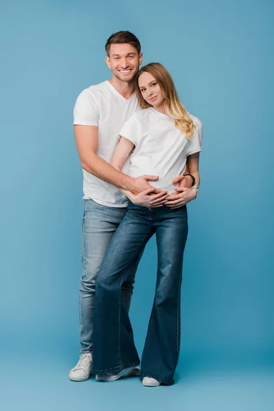 Marito allegro abbracciare sorridente moglie incinta su blu — Foto stock