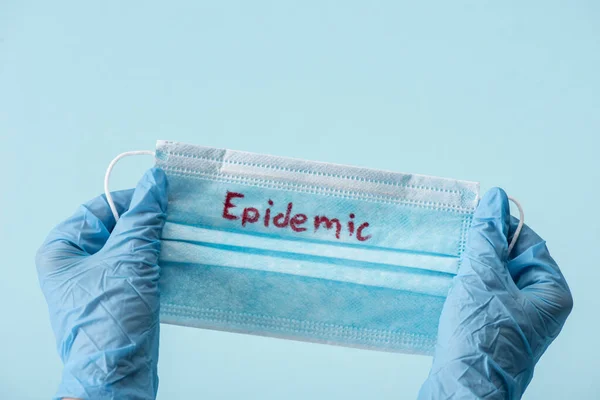 Vista ritagliata del medico in guanti di lattice con maschera medica con iscrizione epidemica isolata su blu — Foto stock