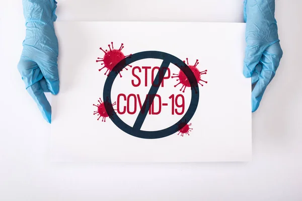 Vue du dessus du médecin en gants de latex tenant une pancarte avec lettrage stop covid-19 sur blanc — Photo de stock