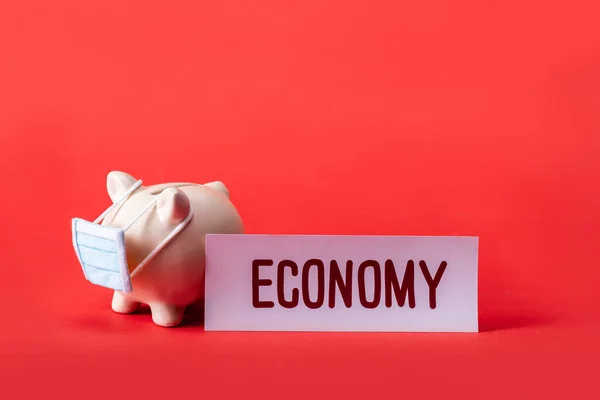 Banco porquinho em pequena máscara médica perto do cartão com letras de economia no vermelho — Fotografia de Stock