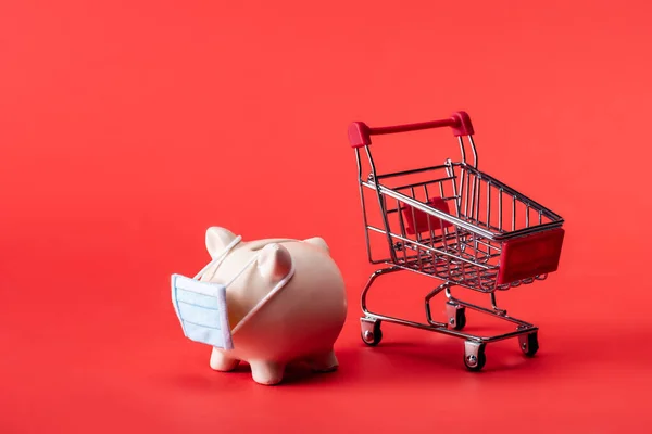Piggy bank in small medical mask near toy shopping cart on red — Stock Photo
