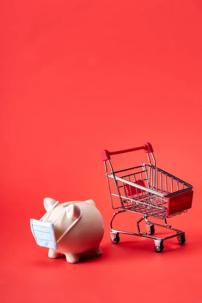 Piggy bank in small medical mask near toy shopping trolley on red — Stock Photo