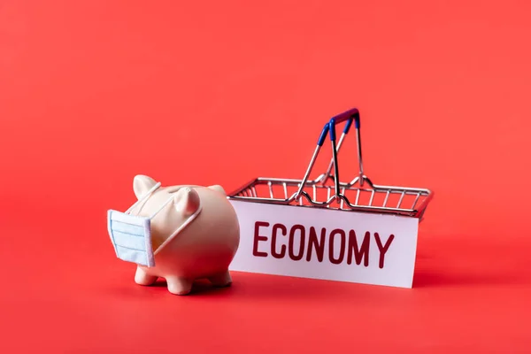 Piggy bank in small medical mask near toy shopping basket and card with economy lettering on red — Stock Photo