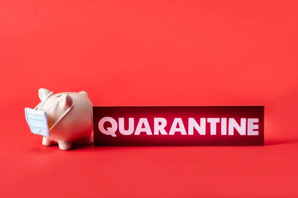 Piggy bank in small medical mask near card with quarantine lettering on red — Stock Photo