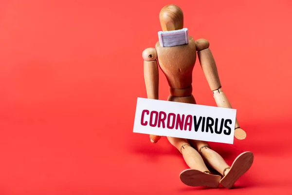 Mannequin en bois en petit masque médical et carte avec lettrage coronavirus sur fond rouge — Photo de stock