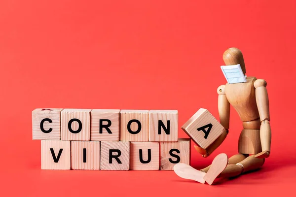 Wooden doll in small medical mask near cubes with coronavirus lettering on red — Stock Photo