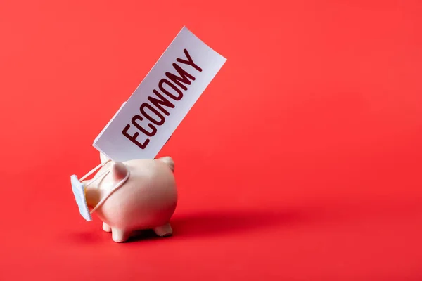 Banco porquinho em máscara médica perto do cartão com letras de economia no vermelho — Fotografia de Stock