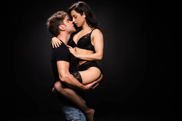 Petit ami passionné tenant copine sexy en lingerie noire sur les mains isolées sur noir — Photo de stock