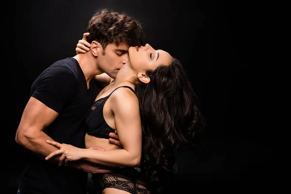 Bell'uomo che abbraccia e bacia ragazza sexy in lingerie nera isolata su nero — Foto stock