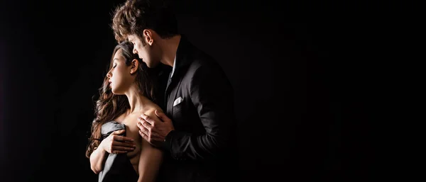 Panoramic shot of handsome man in suit hugging sensual nude woman in black cloth isolated on black — Stock Photo