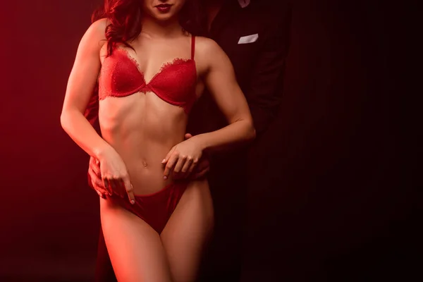 Recortado vista de hombre en traje abrazando sexy chica en lencería roja en negro con luz roja - foto de stock