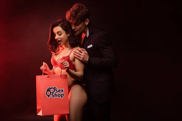 Homem abraçando mulher animada com saco de compras de sex shop em preto com luz vermelha — Fotografia de Stock
