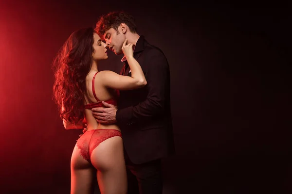 Homme en costume câlin et aller embrasser copine sexy en lingerie rouge sur noir avec lumière rouge — Photo de stock