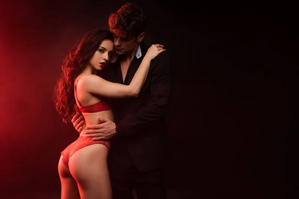 Hombre de traje abrazando novia sexy en lencería roja en negro con luz roja - foto de stock