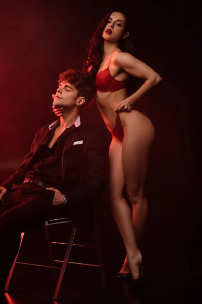 Novia apasionada en lencería roja abrazando novio sexy en traje en negro con luz roja - foto de stock