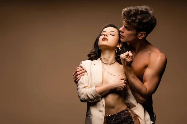 Apasionado novio sin camisa sosteniendo collar de novia medio desnuda seductora en chaqueta beige en beige - foto de stock