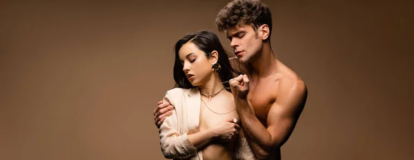 Panoramic shot of passionate shirtless man holding necklace of sexy half naked woman in beige jacket on beige — Stock Photo