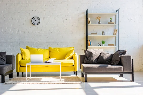 Living room with yellow sofa, grey armchairs, shelf, clock and laptop in sunlight — Stock Photo