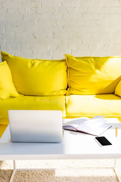 Sala de estar con sofá amarillo y mesa con ordenador portátil, smartphone y bloc de notas a la luz del sol - foto de stock