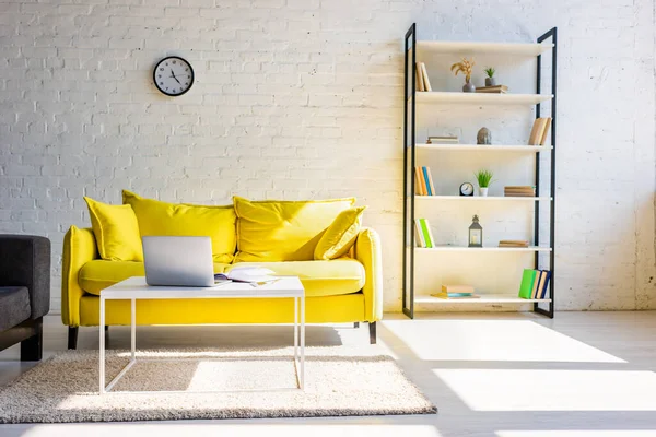 Living room with yellow sofa, shelf and table with laptop in sunlight — Stock Photo