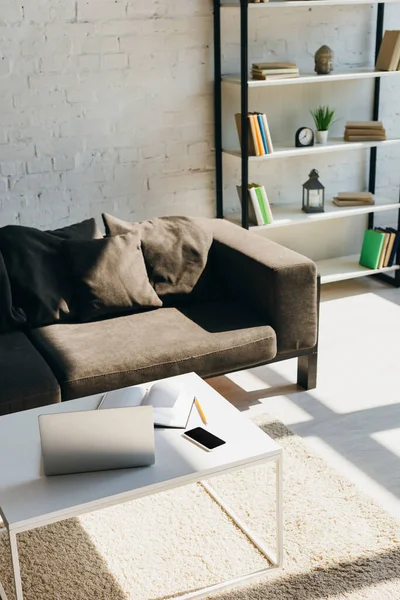 Salon avec canapé gris, étagère et table avec ordinateur portable, smartphone et bloc-notes à la lumière du soleil — Photo de stock