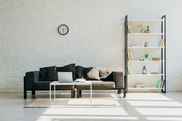Гостиная с серым диваном, часами, полкой и столом с ноутбуком в солнечном свете — стоковое фото