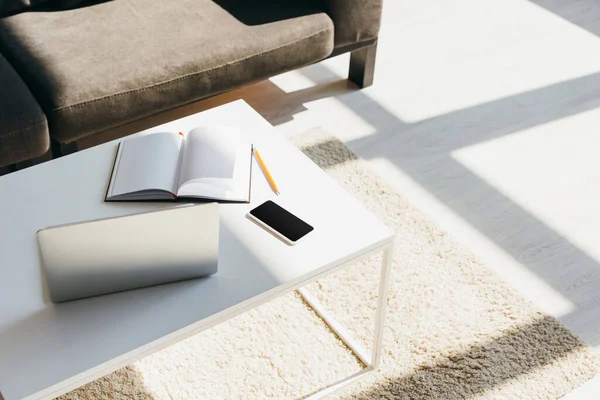 Salón con sofá y mesa con ordenador portátil, smartphone, bloc de notas y lápiz a la luz del sol - foto de stock