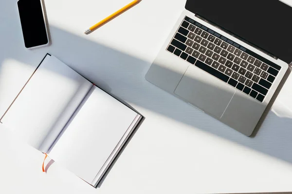 Vista superior do local de trabalho vazio com laptop, smartphone, bloco de notas e lápis na luz solar — Fotografia de Stock