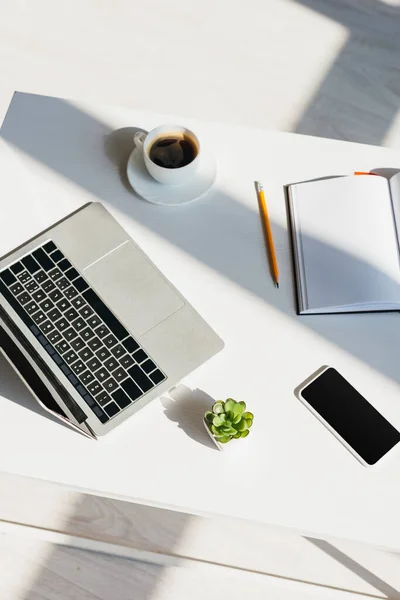 Lugar de trabajo con ordenador portátil, smartphone, bloc de notas y taza de café a la luz del sol - foto de stock