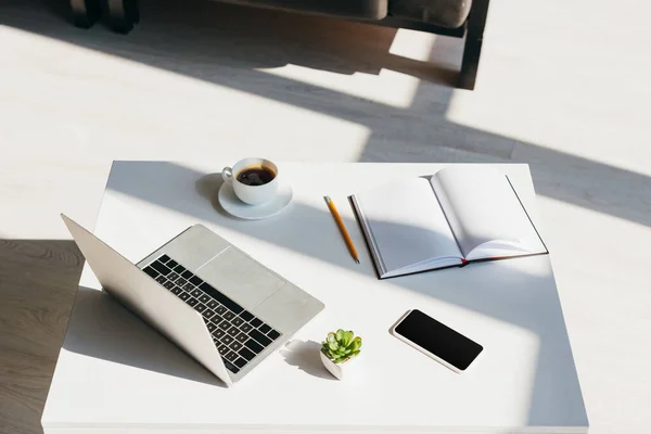 Пустое рабочее место с ноутбуком, смартфоном, блокнотом и чашкой кофе в солнечном свете — стоковое фото