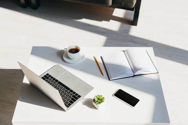 Lugar de trabajo vacío con computadora portátil, teléfono inteligente, bloc de notas y taza de café a la luz del sol - foto de stock