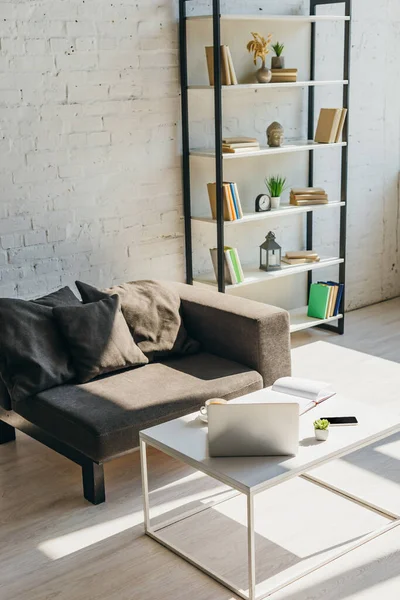 Salon avec canapé gris, étagère et table avec ordinateur portable, smartphone et bloc-notes à la lumière du soleil — Photo de stock