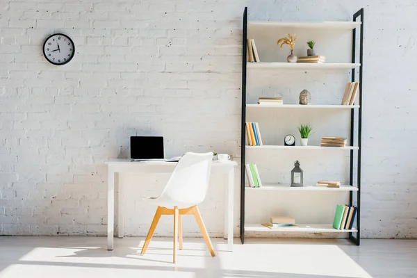 Oficina en casa con estante de libros, reloj, silla y portátil en la mesa a la luz del sol - foto de stock
