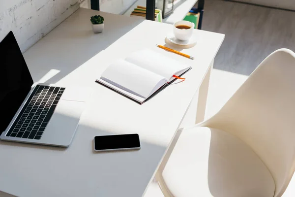 Oficina en casa con teléfono inteligente, bloc de notas, taza de café y computadora portátil en la mesa a la luz del sol - foto de stock