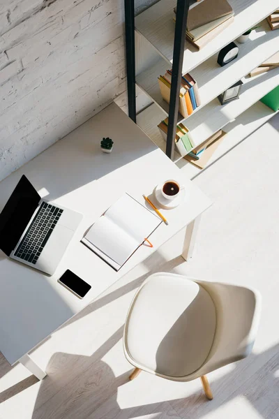 Vista superior de la oficina en casa con ordenador portátil, teléfono inteligente y taza de café a la luz del sol - foto de stock