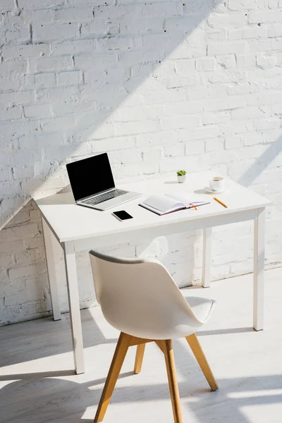 Oficina en casa con ordenador portátil, teléfono inteligente, café y silla a la luz del sol - foto de stock
