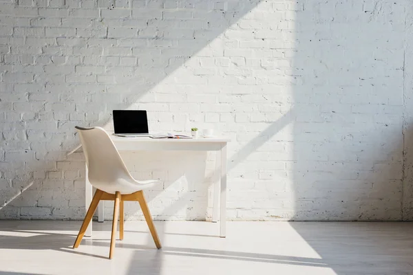 Oficina en casa con mesa, silla y portátil con pantalla en blanco a la luz del sol - foto de stock