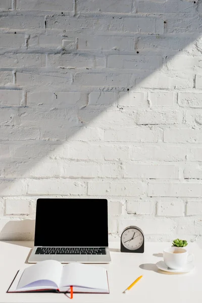 Oficina en casa con ordenador portátil, bloc de notas vacío, reloj y café a la luz del sol - foto de stock