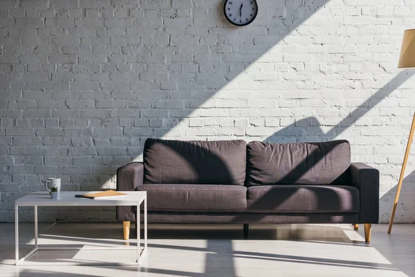 Sala de estar con sofá gris y mesa con taza de café, bloc de notas y suculenta a la luz del sol - foto de stock