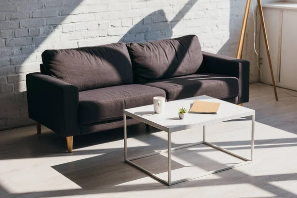 Salon avec canapé gris et table avec tasse de café, bloc-notes et plante de la maison en plein soleil — Photo de stock