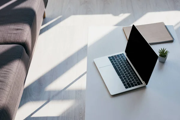 Sofá e mesa com laptop com tela em branco, bloco de notas e planta da casa na luz solar — Fotografia de Stock