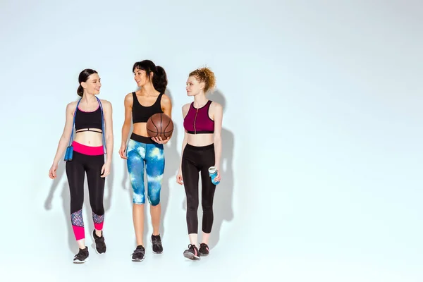 Chica afroamericana sosteniendo baloncesto y caminando con mujeres deportivas en blanco - foto de stock